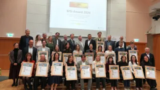 A group of people in formal attire, holding certificates, posing for a photo with a projector screen showing BISi-Award 2026. They are smiling and appear to be winners of the award.