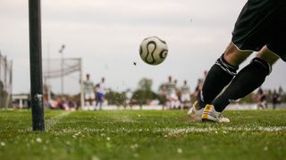 A soccer ball is in midair during a game, with players and spectators in the background.