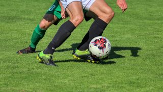 Two soccer players are playing on a grass field. One player is wearing a green jersey and the other is wearing a white jersey. The player in white is kicking the ball.