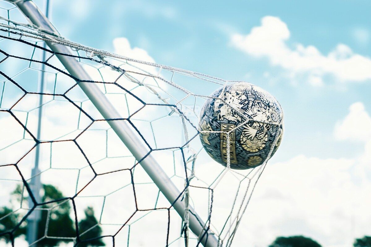 A soccer ball with intricate designs rests in a goal net against a blue sky with clouds.