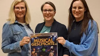 Three women pose for a photo, holding a poster for a musical event on April 17. They are smiling, and the poster has images of characters from the musical.