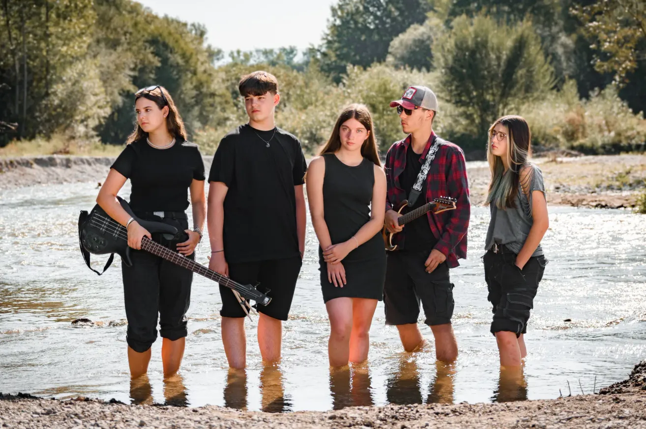 A group of young people stand in a river with guitars, dressed in black and gray. Trees and greenery are in the background.