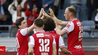 A group of young soccer players in red and white uniforms are celebrating with their hands together. Player number 32 is looking to the side.