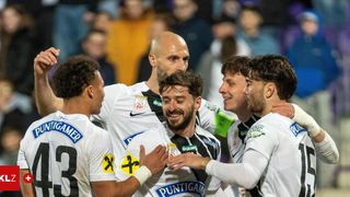 A group of soccer players celebrates a goal, with the central player wearing a shirt with 'Puntigamer' and 'GRAVNE'. Other players wear jerseys with various numbers and brands. Spectators in the background watch.