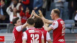 Soccer players celebrate a goal with hands high in the air in front of an excited crowd. The team wears red jerseys with white numbers.