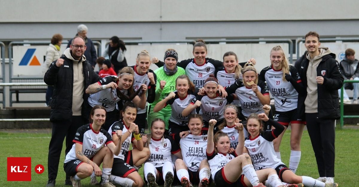 A women's soccer team poses for a photo with their arms raised in celebration, wearing white uniforms with numbers on the back. The background shows a building and a fence.