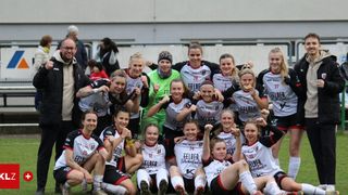 A women's soccer team poses for a photo with their arms raised in celebration, wearing white uniforms with numbers on the back. The background shows a building and a fence.