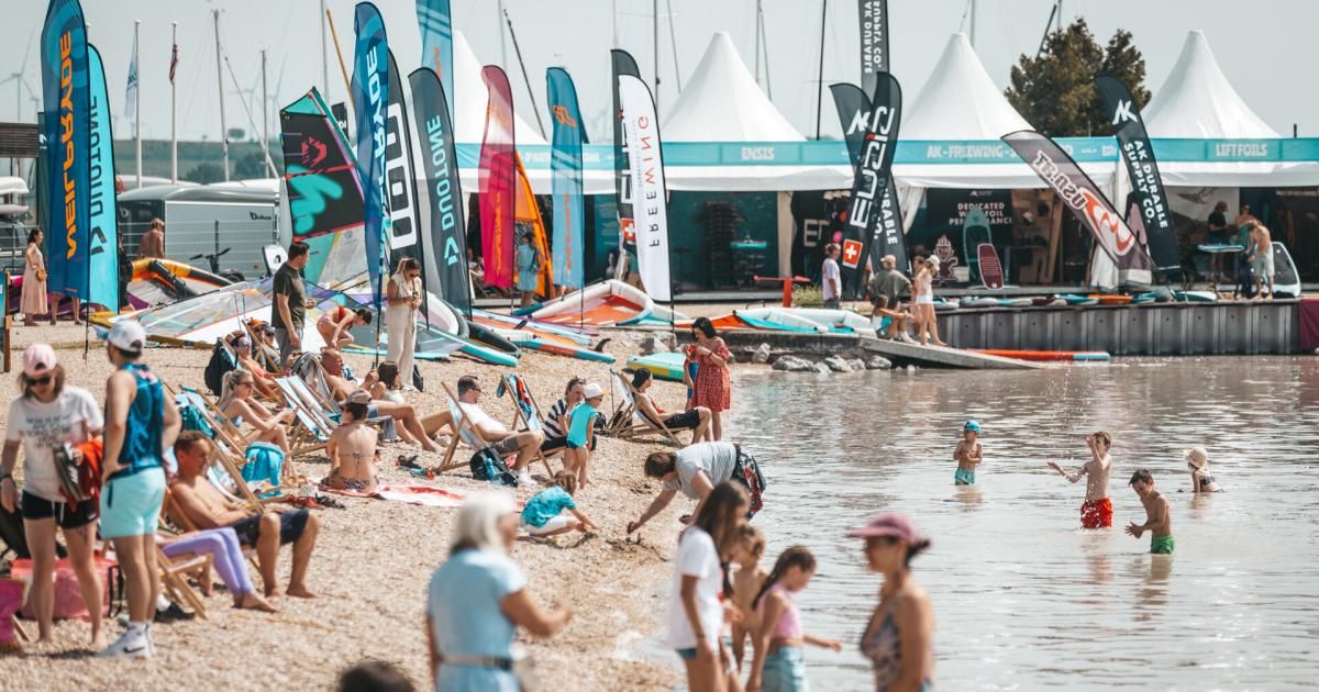A beach filled with people, some sitting on chairs, others standing in water. Banners are visible, and some people are under tents. The water is calm.