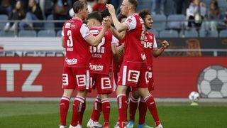 Soccer players in red uniforms celebrate a goal on the field, with red banners and spectators in the background.