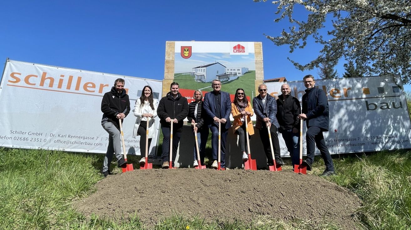 Eine Gruppe von Menschen steht vor einer Baustelle, hält Schaufeln und posiert für ein Foto. Im Hintergrund befindet sich ein großes Schild mit einem Bild eines Hauses.