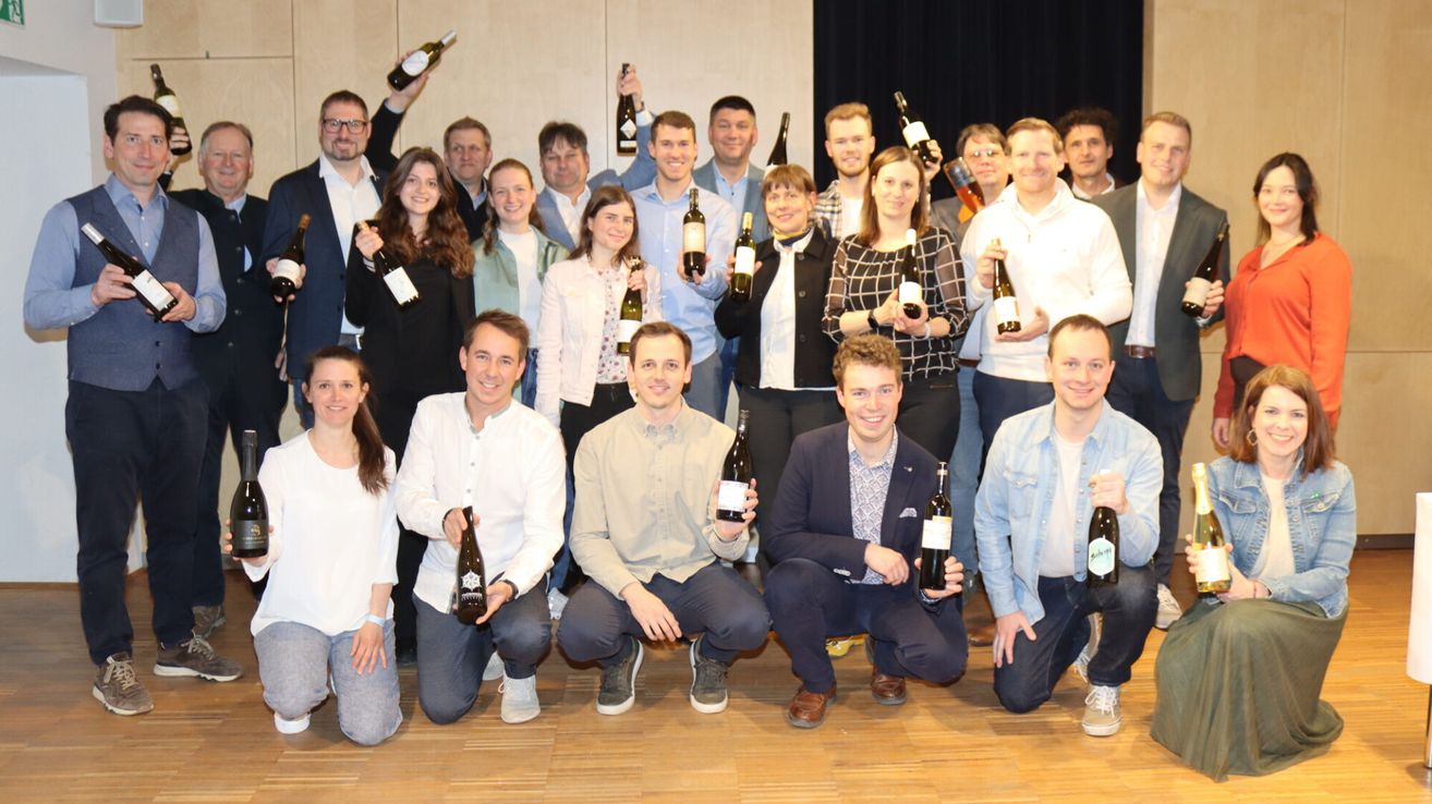 A group of people are posing for a photo with wine bottles in their hands. They are all smiling and some are holding wine bottles. They are probably at a wine tasting event.