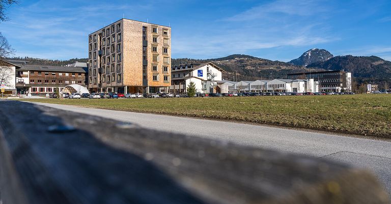 A tall building with many windows stands behind a grassy area. Several cars are parked in front of the building. In the distance, there are mountains and other buildings.