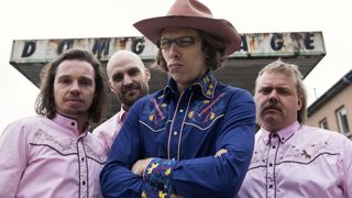 Four men in pink shirts and hats stand together. The central man wears a blue shirt with guitars on it and has a cowboy hat.