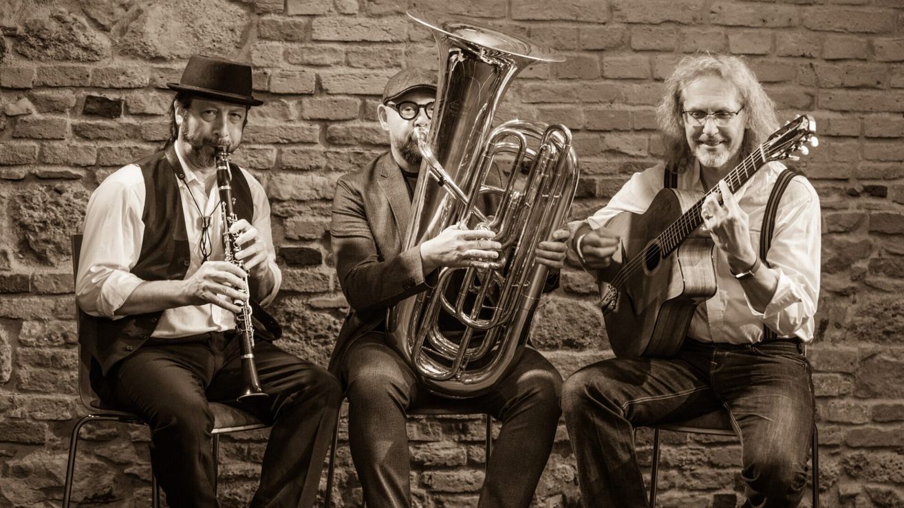 Three musicians are seated in front of a brick wall, each playing a different instrument: a tuba, a clarinet, and a guitar. They are dressed in formal attire and appear to be performing.