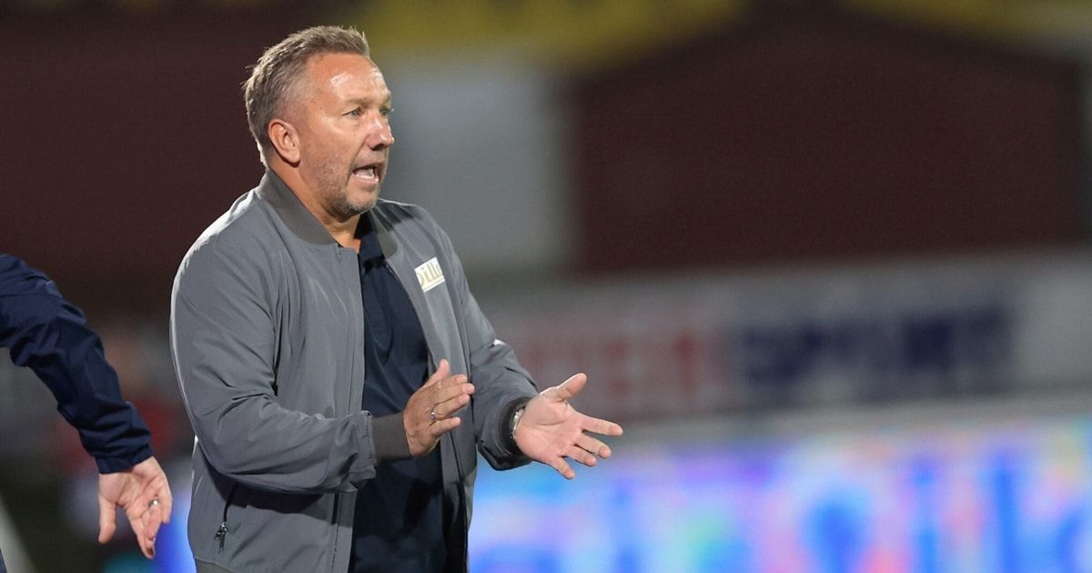 A man in a gray jacket with the word 'DIL' printed on it is speaking on a field. He appears to be a coach.