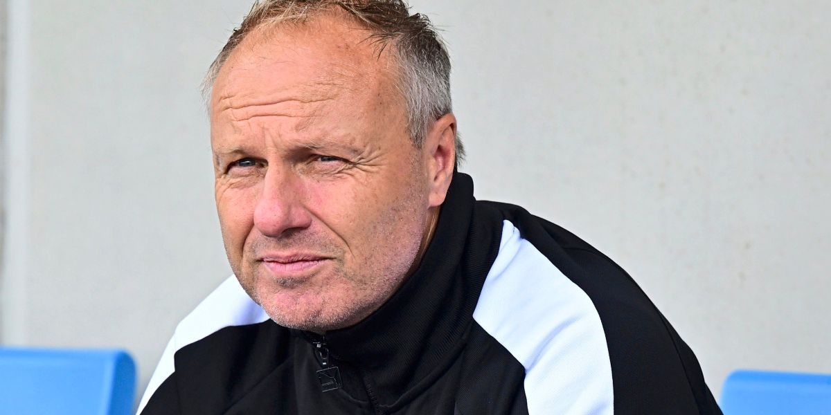 A man in a black and white jacket with a zipper looks serious and slightly sad. He has a white beard and is standing in front of a white wall.