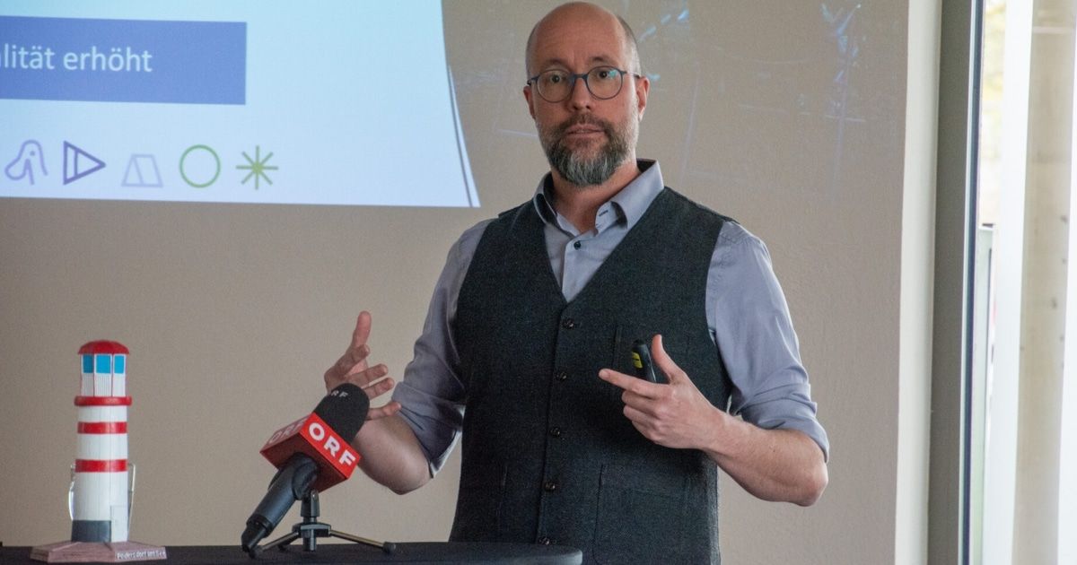 A man stands at a podium with a microphone, wearing a vest and glasses. Behind him is a projector screen displaying a blue square with a white star.