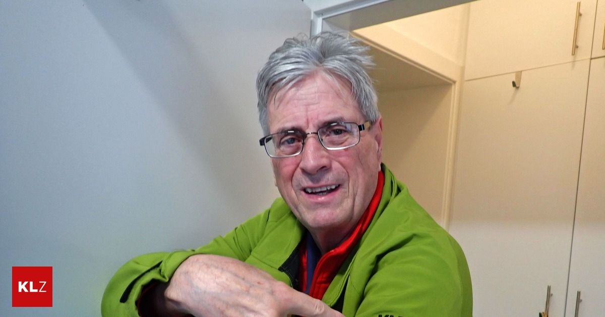 A man wearing a green jacket with a red collar points at something with his right hand while smiling.