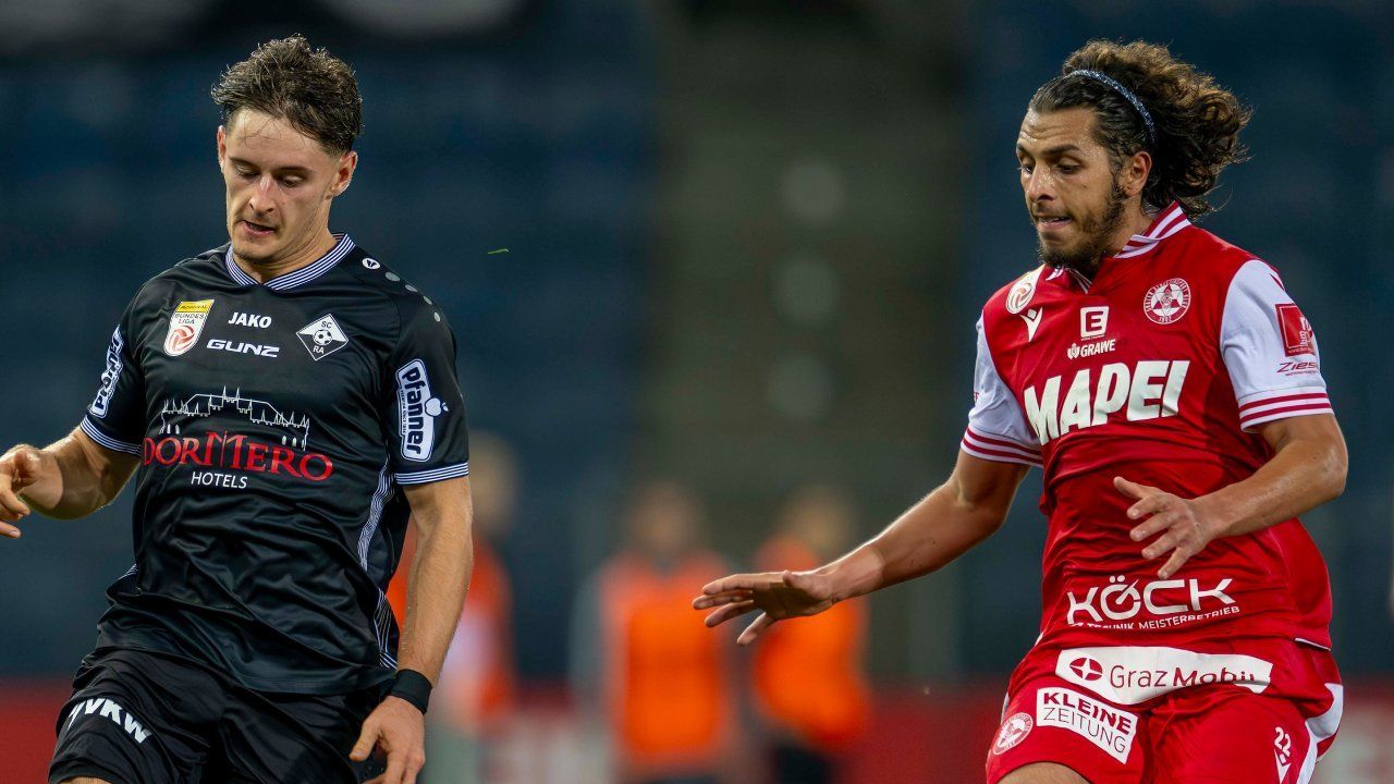 Two soccer players in action. One wears black, the other red, both focused. They are likely in a competitive match.