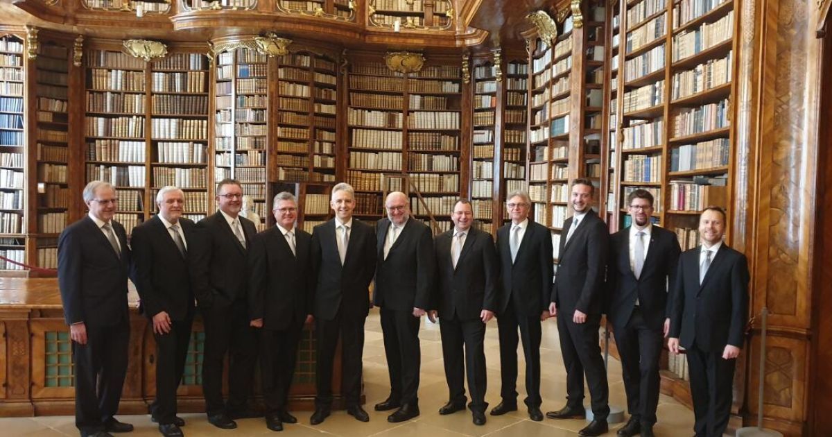 A group of formally dressed men stand in a library, posing for a photo among rows of bookshelves.