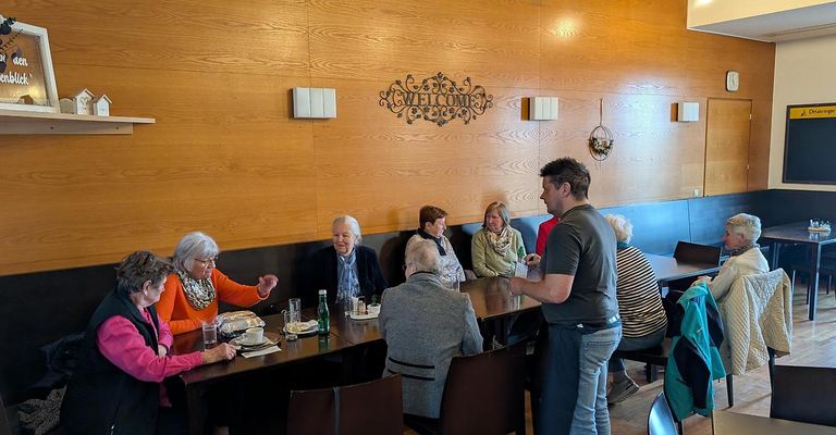 A group of elderly women and a man sit around a table in a restaurant, engaged in conversation. The wooden wall has a 'Welcome' sign, and a man stands nearby.
