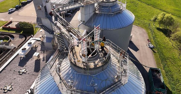 A top-down view of a large industrial facility with two massive cylindrical storage tanks, workers on a ladder and on platforms, and surrounding stairs and pipes.