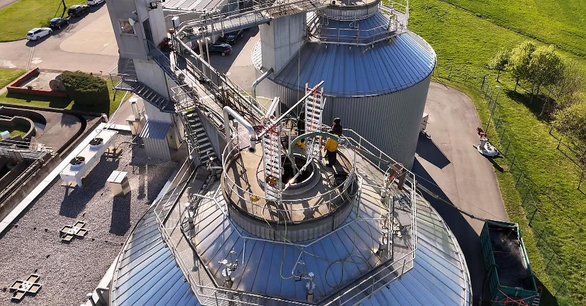 A top-down view of a large industrial facility with two massive cylindrical storage tanks, workers on a ladder and on platforms, and surrounding stairs and pipes.