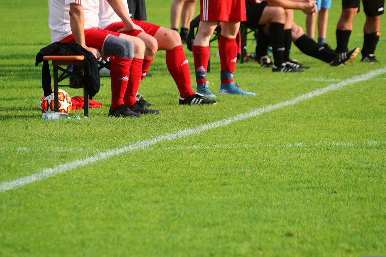 Soccer players sit on the bench, wearing red uniforms, socks, and sneakers. Some wear knee pads, and a soccer ball rests nearby. White lines mark the field.