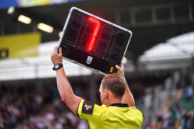 Ein Fußballschiedsrichter hält eine rote elektronische Karte mit der Nummer 7. Der Mann trägt ein gelbes Trikot und steht vor einem unscharfen Publikum.