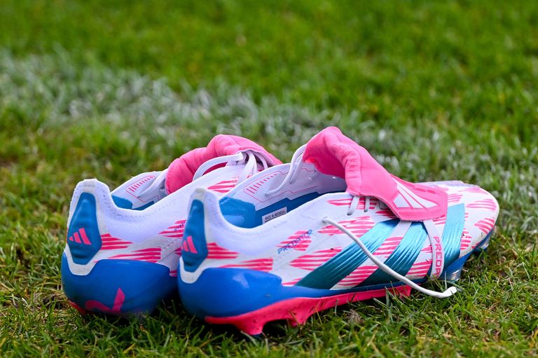 A pair of soccer shoes with pink, blue, and white colors are on the grass. They have a brand name 'PREDATOR'. The cleats have a white sole.