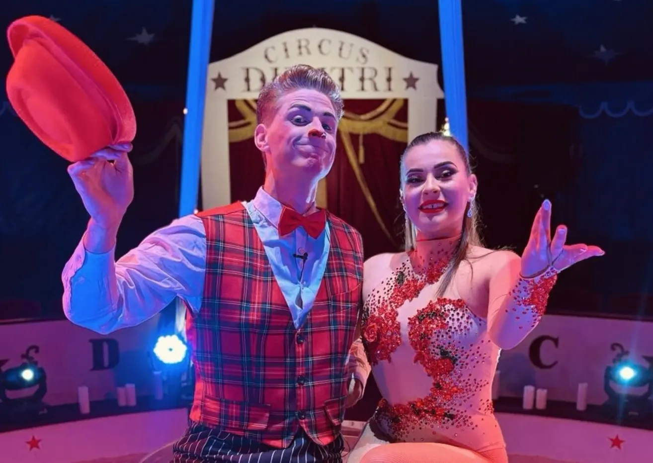 A man and a woman dressed in circus costumes, smiling and posing for a photo in front of a Circus Dutri banner.
