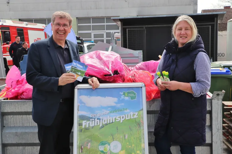 A man and woman stand next to a sign advertising a spring event, holding brochures and a small item, with bags in the background.