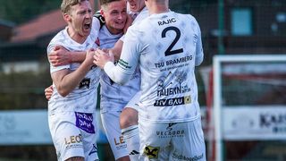 Four young soccer players in white jerseys celebrate a victory. The player in the middle wears a jersey with number 2. They are smiling and hugging each other.