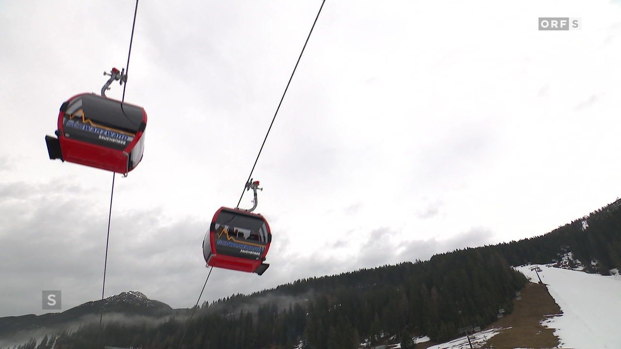A red cable car with passengers is suspended from a cable in a mountainous region, amidst a cloudy sky. The cable car features a logo on its side.