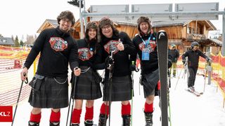 Four people in kilts stand on a snowy slope, smiling. They wear ski boots and carry ski poles. A wooden building and a ski lift are in the background.