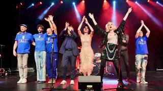 A group of musicians are performing on a stage, with a woman singing and raising her hands, and a man in the background.