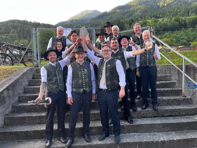 A group of men in traditional clothing pose for a photo on steps, holding a trophy and a glass, with mountains in the background.