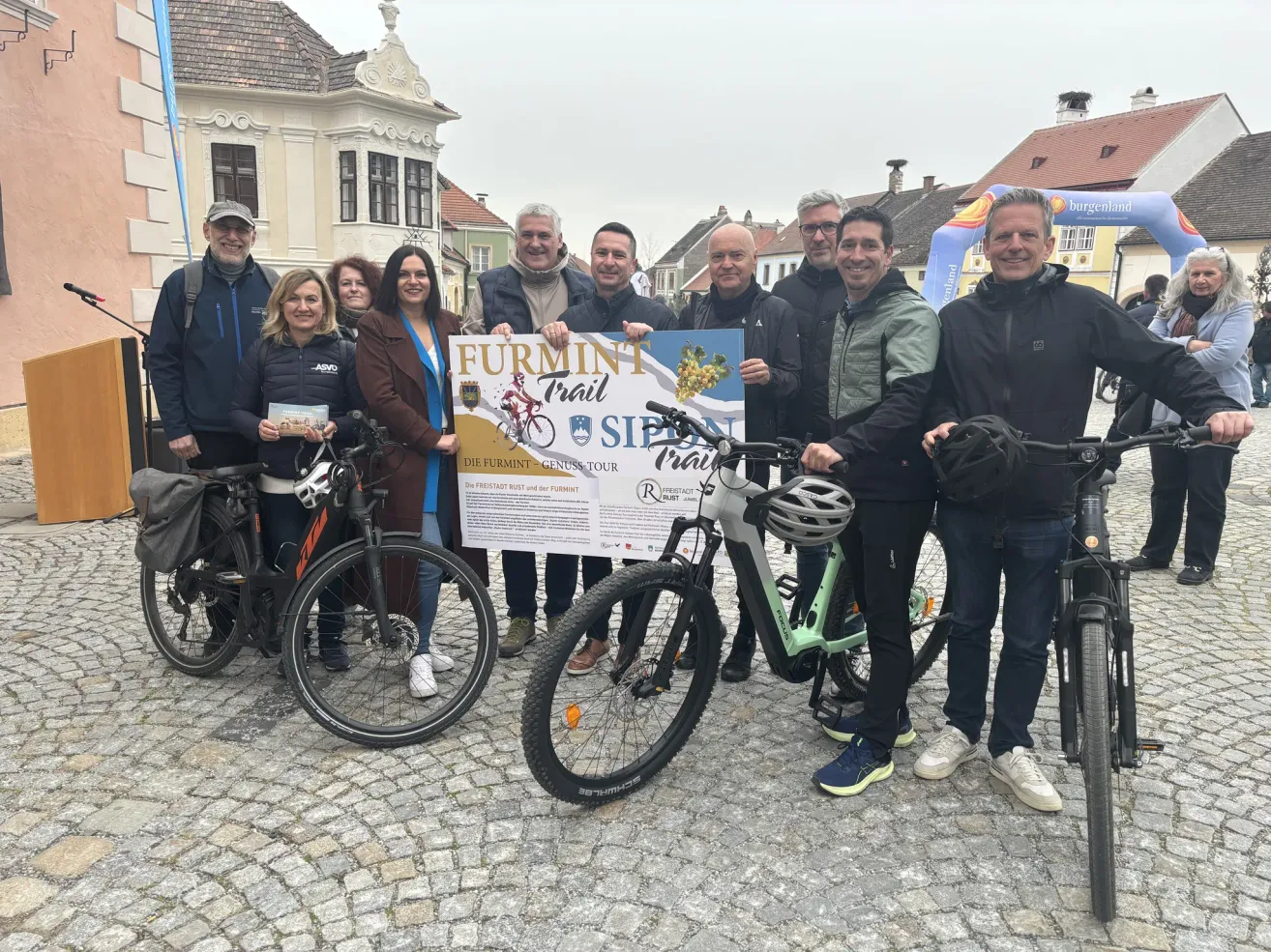 A group of people pose for a photo with bicycles and a banner. They are in front of buildings and cobblestone streets.