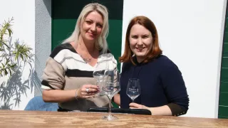Two women smile and hold wine glasses with labels reading 'PURBACH' and 'PURBACH 1993'. They are seated at a table, with a green door behind them.