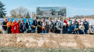 A group of people at a construction site holding shovels, with a large sign in the background reading 'Mittelschule Seiersberg Fertigstellung Sommer 2027'.