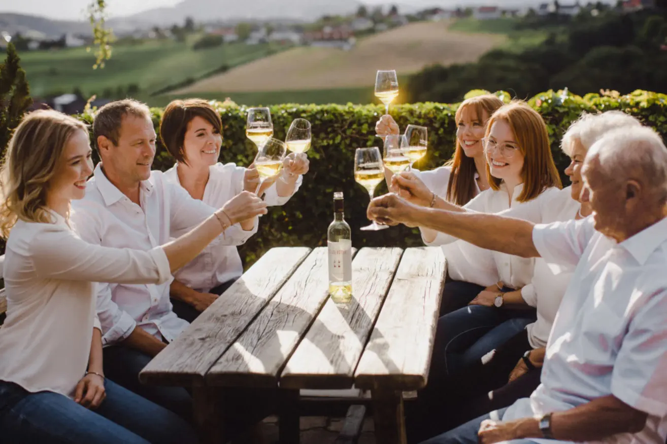 Five people, smiling and holding wine glasses, are sitting around a wooden table, possibly celebrating a special occasion. A bottle of wine is on the table. Behind them is a green hedge, with a view of a vast field and mountains in the distance.
