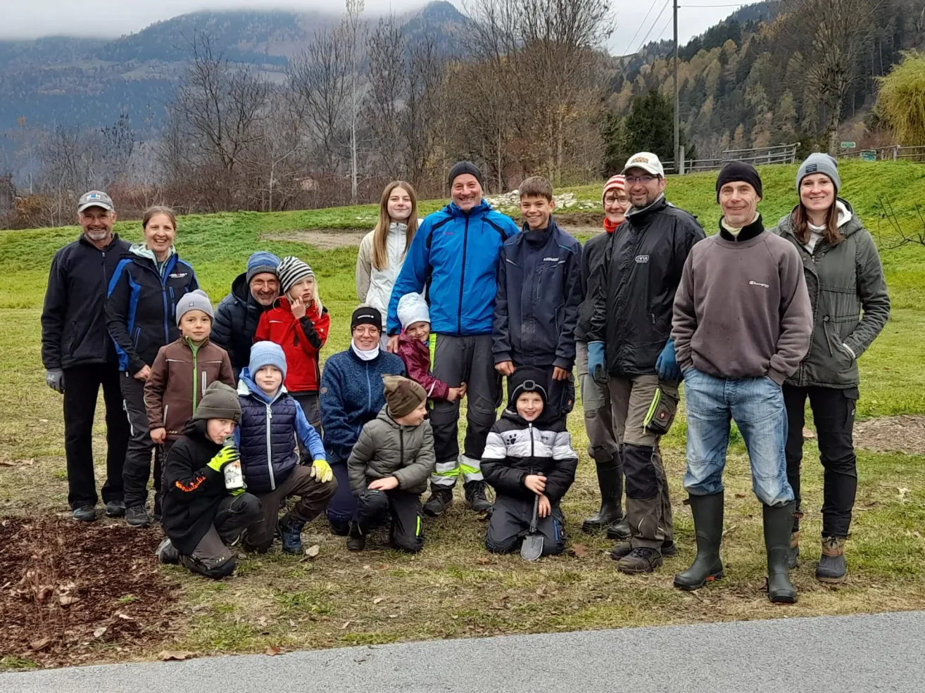 A group of people are standing and sitting on a grassy field, probably posing for a photo. They are wearing winter clothes. One of them is holding a shovel. Behind them is a road with a pathway. In the distance are mountains and trees.