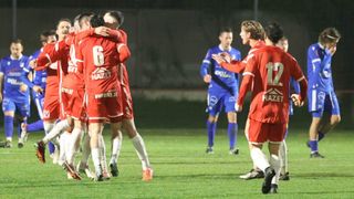 A group of soccer players in red jerseys celebrate on the field at night. A player in blue is running.