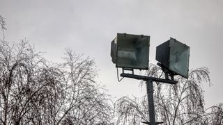 A streetlight with two floodlights is positioned in front of a backdrop of leafless trees under a gray sky.