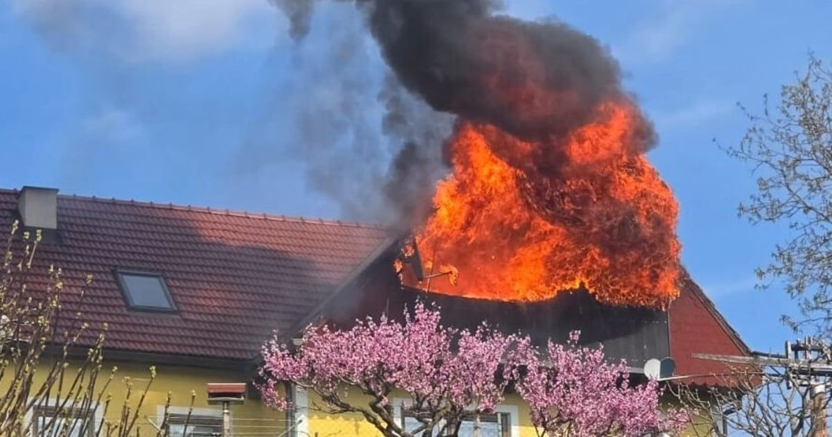 A yellow house with a red roof is on fire. The fire is spreading from the roof to the windows. Black smoke is coming from the flames.