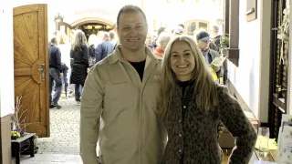 A man and woman stand smiling for a photo in a bustling street, with a tavern sign in the background.