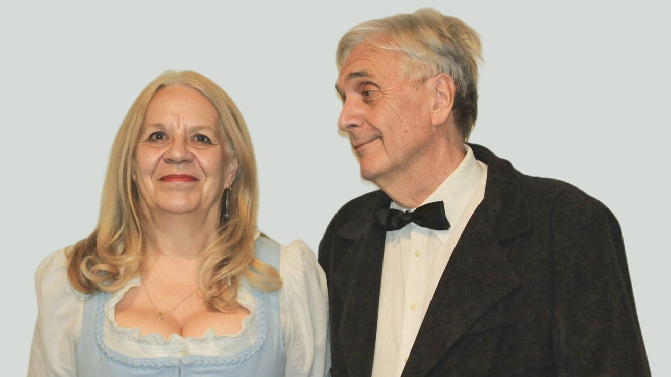 An elderly man and woman stand close together, both smiling. The woman wears a white blouse with a blue bodice and a necklace. The man wears a black tuxedo with a black bowtie.