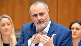 A man in a blue suit speaks into a microphone. He has gray hair and a beard, and is gesturing with his hand.