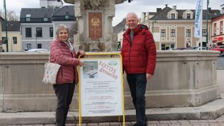 Two individuals, a man and a woman, stand near a sign and a fountain. The sign reads 'Fotos gesucht', and they are dressed warmly in winter clothing. Behind them, a building with a fountain and a plaque is visible.