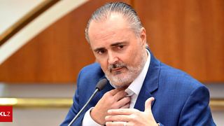 A middle-aged man with a beard is speaking into a microphone. He wears a blue suit and a white shirt. He gestures with his hand while looking forward.